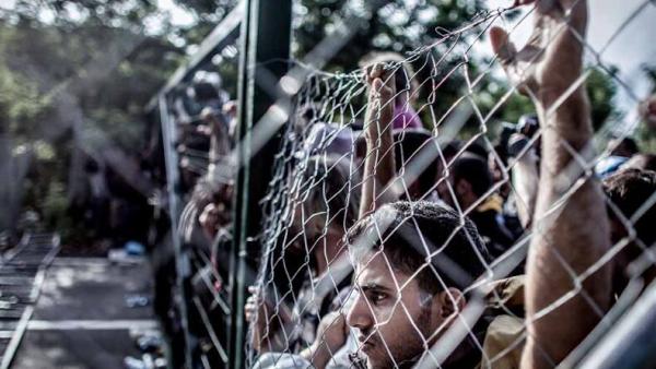 Vários migrantes encostados a uma rede de metal.